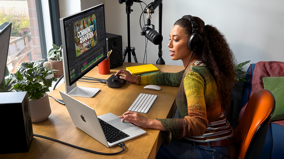 Žena sedí doma u pracovního stolu a používá MacBook Pro připojený k externímu monitoru, má nasazená sluchátka a mluví do mikrofonu