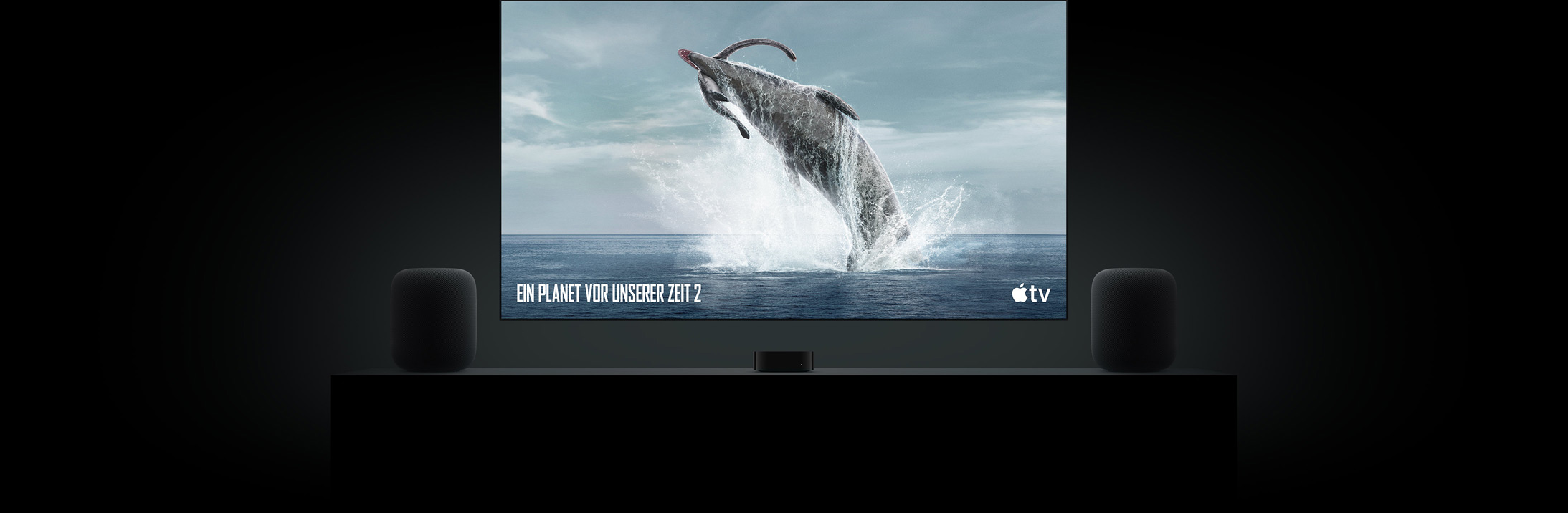 Ein großer Flachbildfernseher zeigt ein brillantes Bild eines Dinosauriers aus Ein Planet vor unserer Zeit. Der Fernseher hängt über einem Apple TV 4K in der Mitte von zwei HomePod Lautsprechern, die auf einem Sideboard in einem Wohnzimmer stehen.