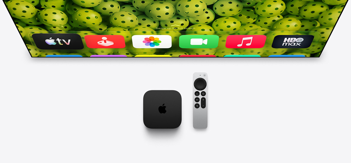 Apple TV 4K og Siri Remote under en TV-skærm, skærmen viser hjemmeskærmen med appsymboler i øverste række og en baggrund med grønne bolde med huller i