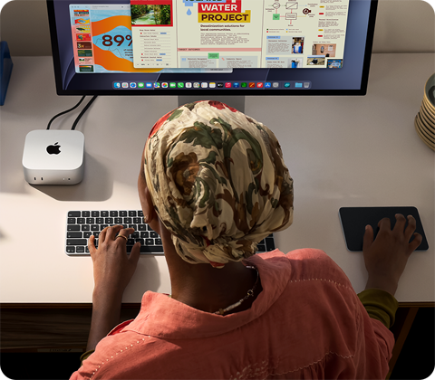 Людина за робочим столом друкує на Mac mini, до якого підʼєднано Studio Display, клавіатура Magic Keyboard, трекпед Magic Trackpad, різноманітні предмети