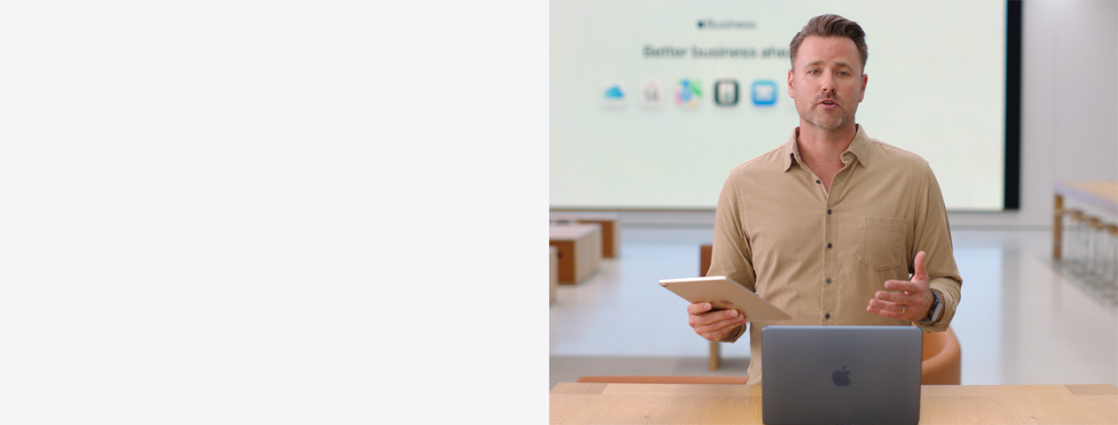 De presentator spreekt met een iPad in de hand en een MacBook voor zich op tafel, terwijl op een groot scherm verschillende symbolen van zakelijke apps te zien zijn.
