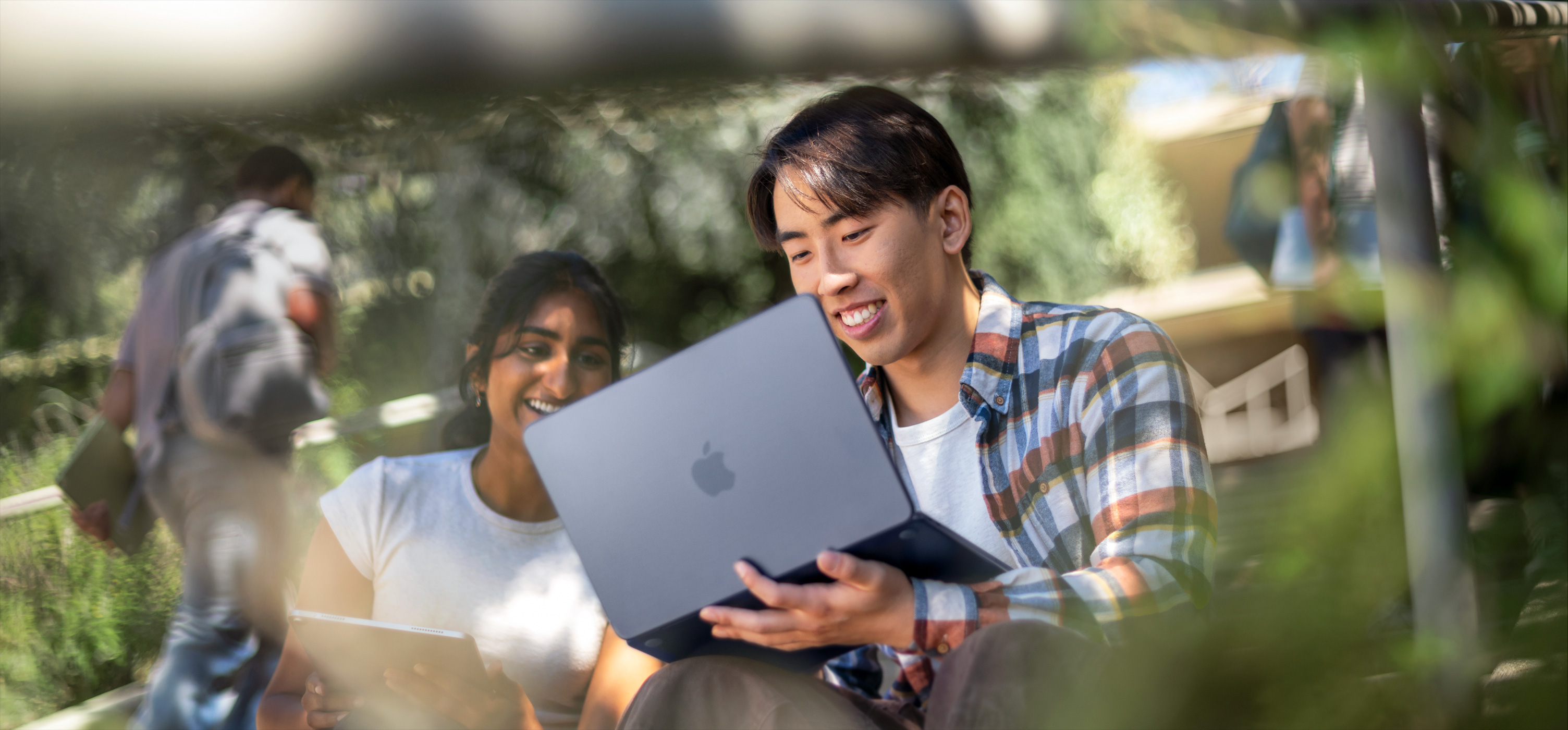 屋外でMacBookを使いながら、笑顔で課題の共同作業に取り組んでいる2人の学生。1人は膝の上にMacBookを置いている