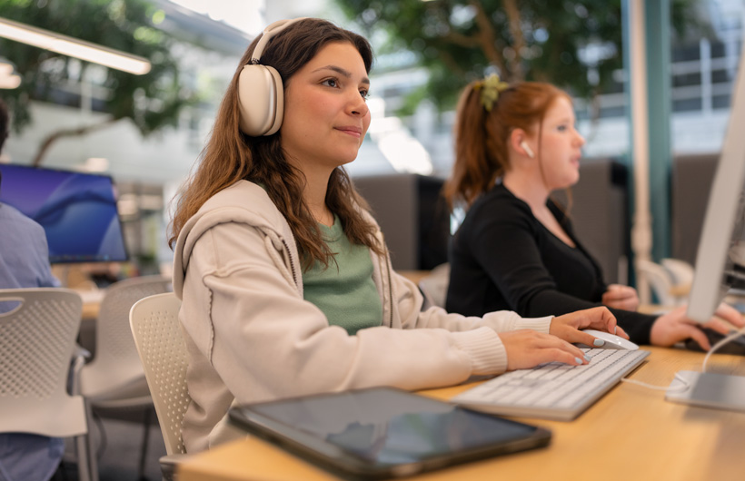 A student wearing AirPods&nbsp;Max sits at a computer typing on a keyboard, with a tablet resting nearby, while another student works next to her.
