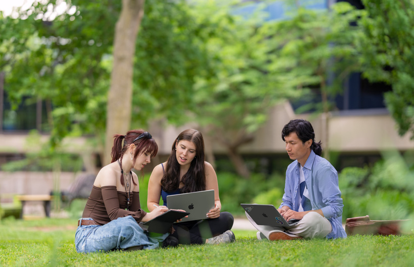 Three students sit in the grass on an outdoor campus, one using a MacBook&nbsp;while another looks on.