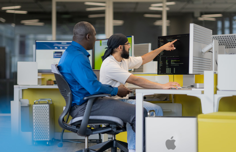 Two students sit in front of multiple iMac&nbsp;monitors, one pointing at a screen.