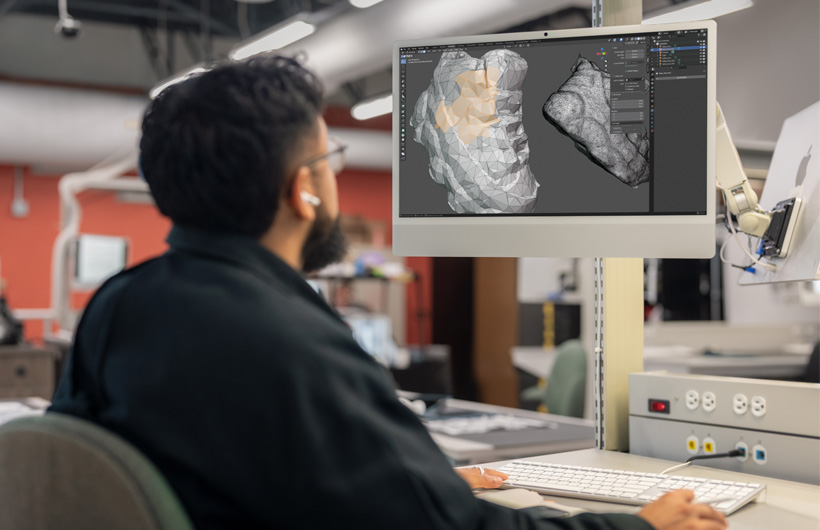 A student sits at a desk reviewing two 3D-scanned head models displayed side by side on iMac&nbsp;monitors.