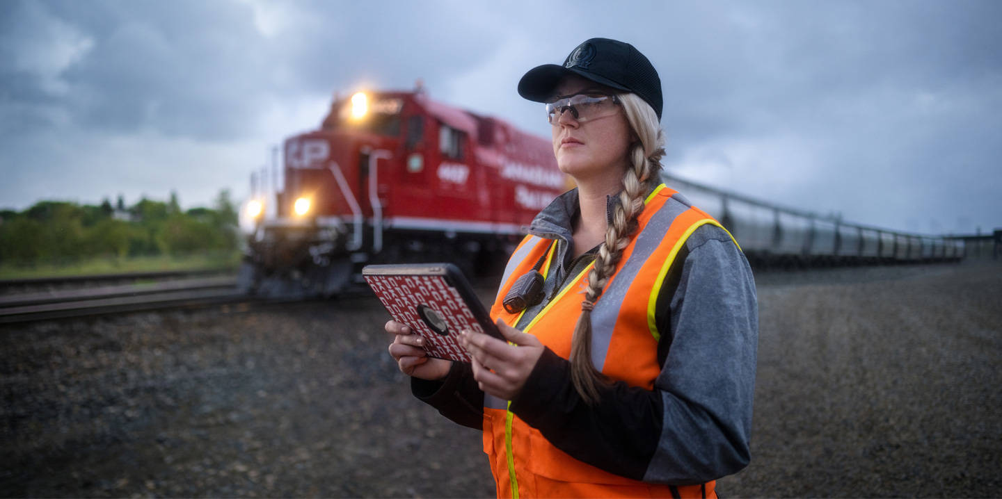 Eine Bahnmitarbeiterin hält ein iPad in der Hand und schaut auf die Gleise, während ein Zug vorbeifährt