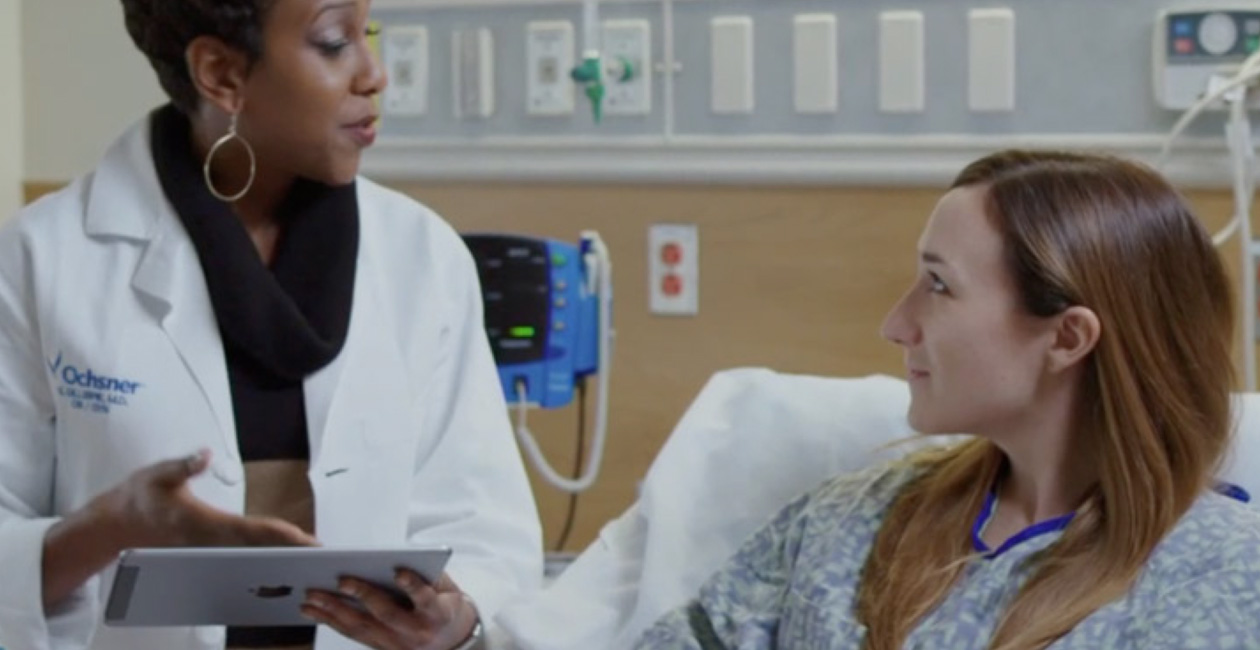 Eine Pflegekraft mit einem iPad spricht mit einer Patientin, die im Krankenhausbett sitzt.