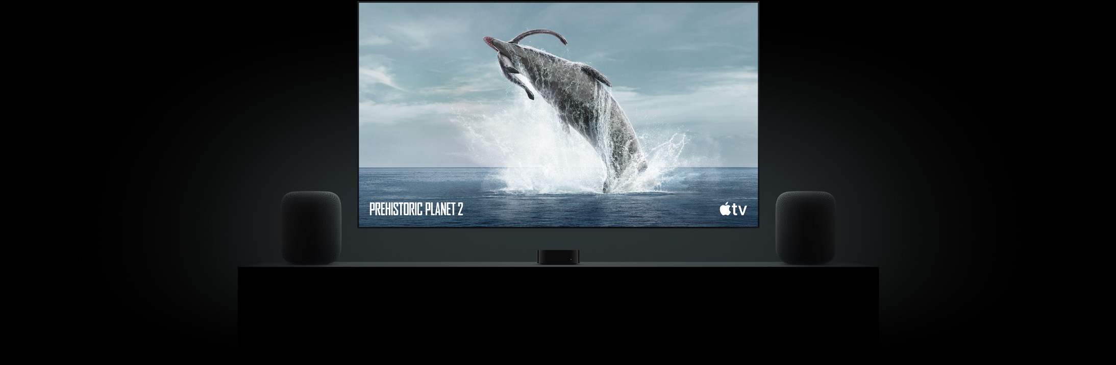 Ein großer Flachbildfernseher zeigt ein brillantes Bild eines Dinosauriers aus Ein Planet vor unserer Zeit. Der Fernseher hängt über einem Apple TV 4K in der Mitte von zwei HomePod Lautsprechern, die auf einem Sideboard in einem Wohnzimmer stehen.